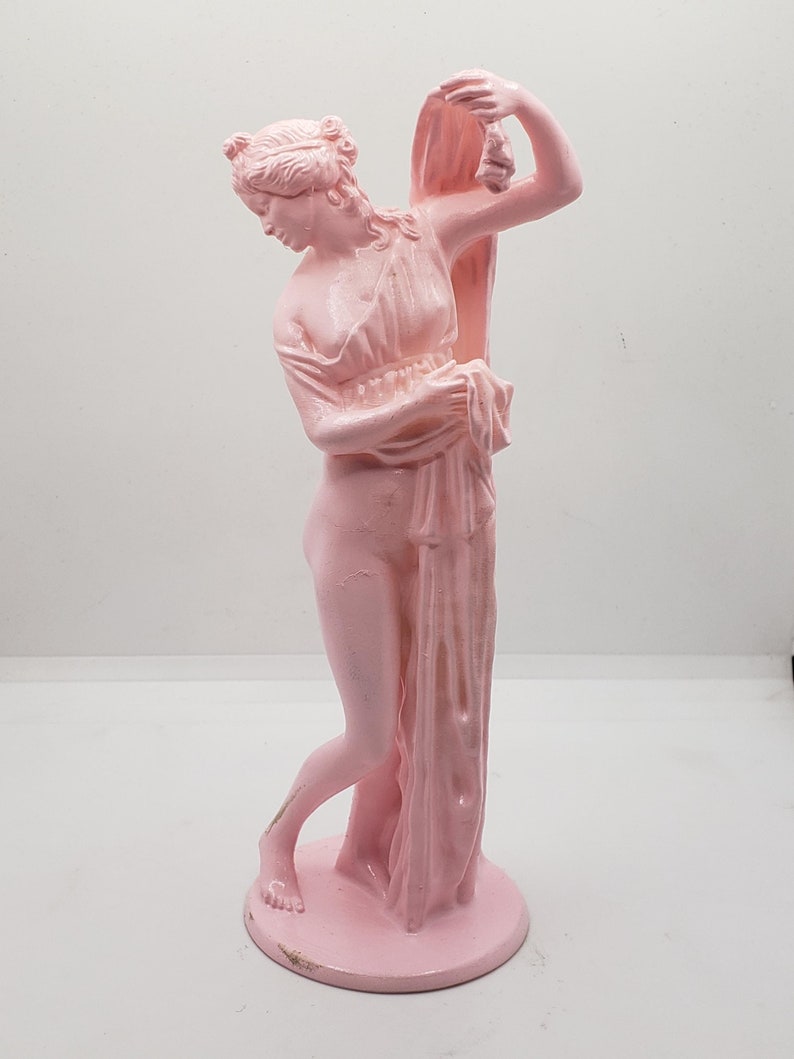 May include: A pink ceramic statue of a woman standing with her arm raised, holding a drape over her head. The statue is on a round base.