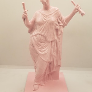 May include: A pink painted statue of a woman holding two scrolls in her hands. The statue is standing on a square base.