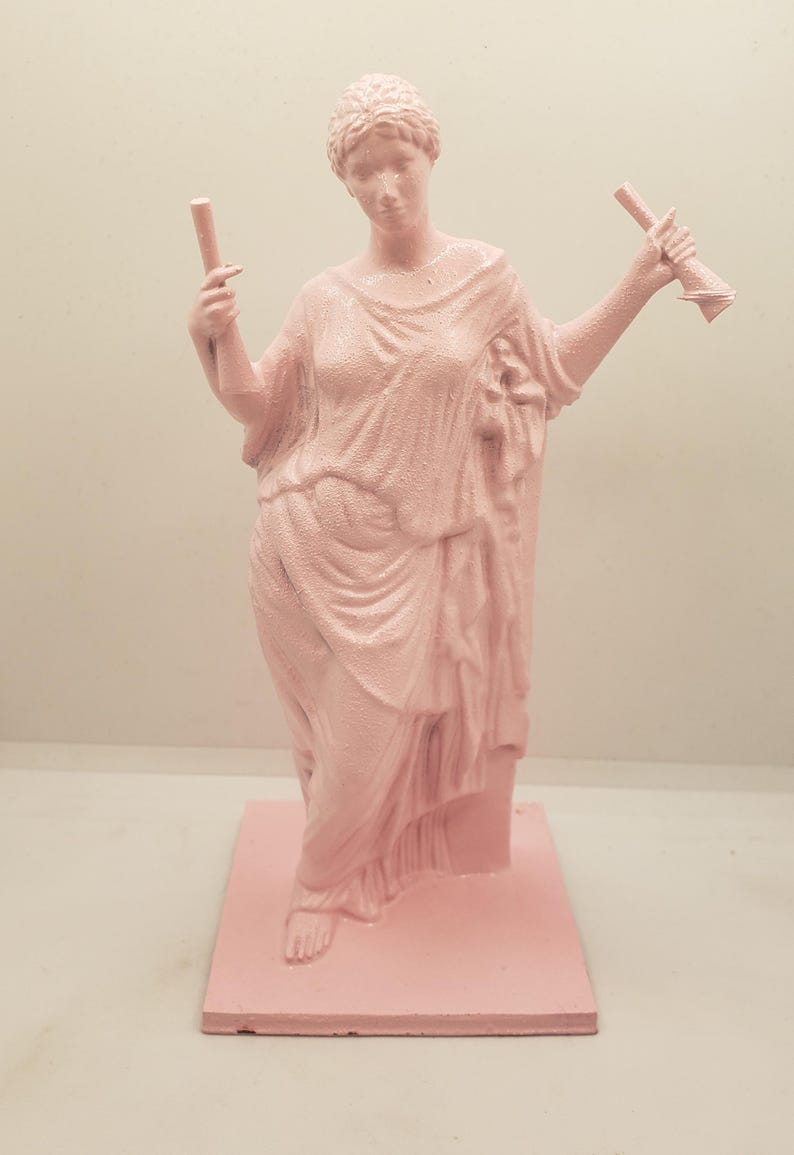 May include: A pink painted statue of a woman holding two scrolls in her hands. The statue is standing on a square base.