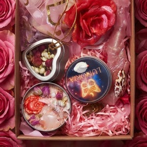 May include: A rectangular cardboard box filled with pink paper shreds, containing a heart-shaped tin with dried flowers, a round tin labeled Lunoria with a butterfly graphic, a red rose-shaped wax item, a pink crystal-like object, and a seashell.