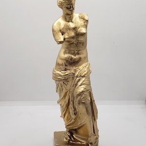 May include: A gold-colored statue of the Venus de Milo, a famous Greek sculpture. The statue is standing on a square base and is missing its arms.