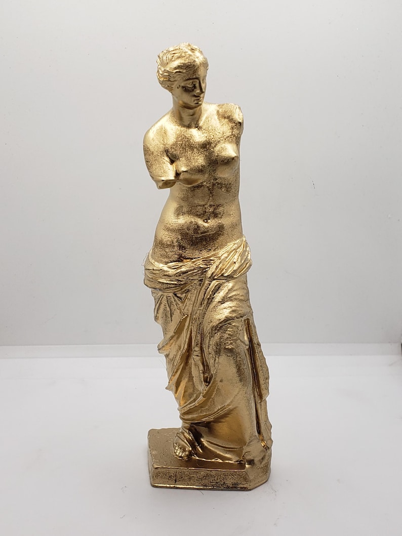 May include: A gold-colored statue of the Venus de Milo, a famous Greek sculpture. The statue is standing on a square base and is missing its arms.