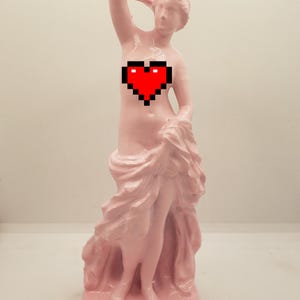 May include: A pink painted statue of a woman with a red pixel heart on her chest. The statue is standing on a pink base and is wearing a flowing pink robe.