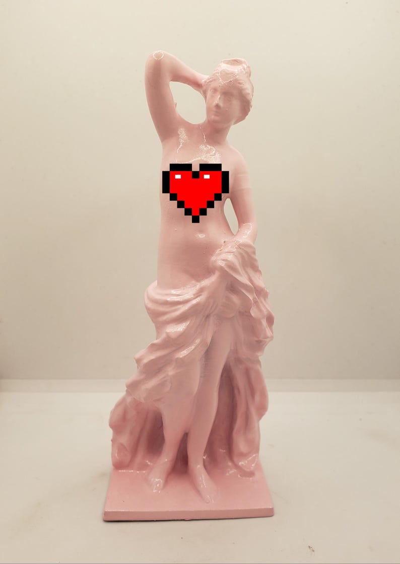 May include: A pink painted statue of a woman with a red pixel heart on her chest. The statue is standing on a pink base and is wearing a flowing pink robe.
