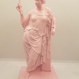 May include: A pink painted statue of a woman in a flowing robe, standing on a square base. The woman is holding a small object in her right hand.