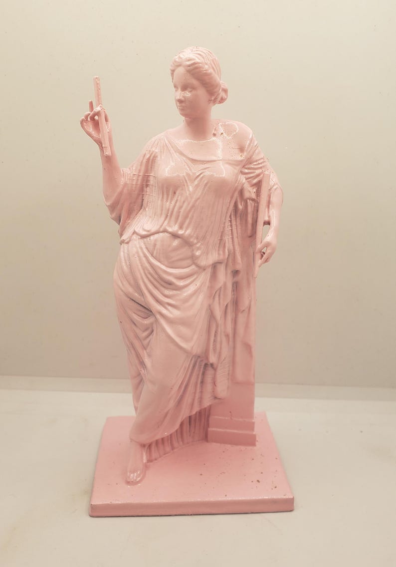 May include: A pink painted statue of a woman in a flowing robe, standing on a square base. The woman is holding a small object in her right hand.