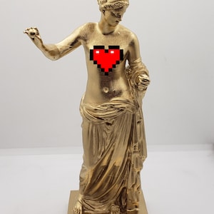 May include: A gold-colored statue of a woman with a pixelated red heart on her chest. The statue is standing on a square base.