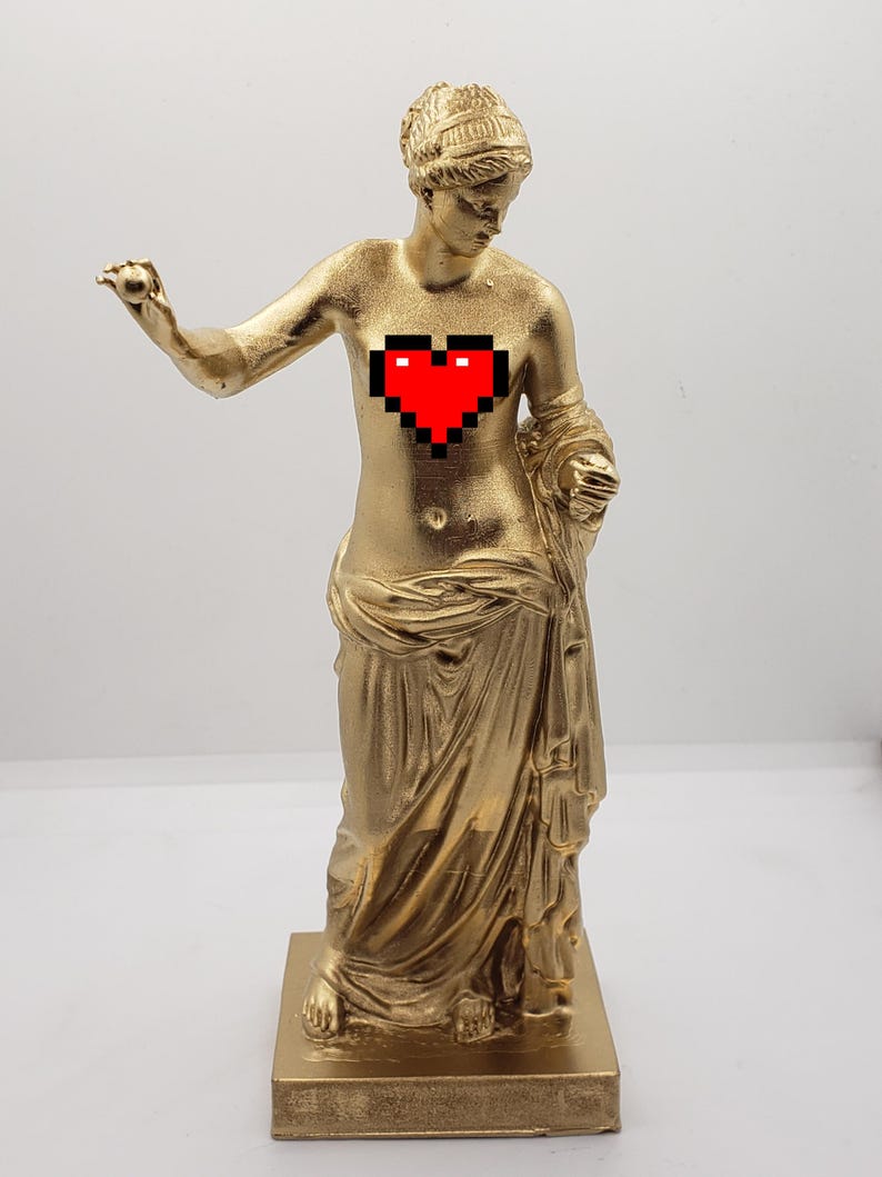 May include: A gold-colored statue of a woman with a pixelated red heart on her chest. The statue is standing on a square base.