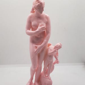 May include: A pink statue of a woman and a child, both nude, standing on a dolphin. The woman is holding her hand to her chest and the child is holding a bow and arrow.