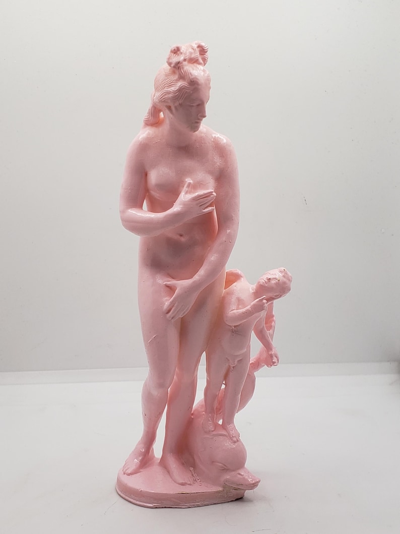 May include: A pink statue of a woman and a child, both nude, standing on a dolphin. The woman is holding her hand to her chest and the child is holding a bow and arrow.