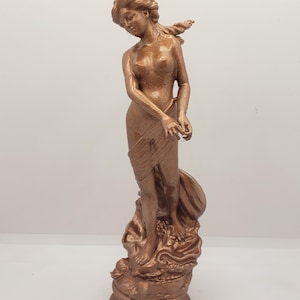 May include: A copper-colored statue of a woman standing on a base with a flowing fabric draped around her. The statue is detailed with intricate carvings and a smooth finish.