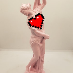 May include: A pink painted statue of a woman holding a drape. The statue is standing on a white surface. A pixelated red heart is covering the woman's chest.
