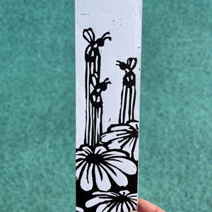 May include: A black and white bookmark featuring a linocut print. The design includes stylized flowers at the bottom, vertical striped figures, and two insect-like figures. The bookmark is held against a teal background.