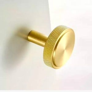 May include: A brass-coloured cabinet knob with a textured, ridged edge and a smooth, circular face. The knob is cylindrical and mounted on a white surface, casting a shadow.