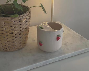 May include: White ceramic mug with a strawberry design. The mug has a unique shape with a cut-out on one side. The mug is sitting on a white marble surface.