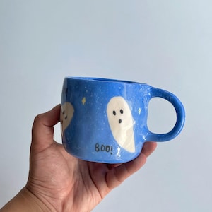 May include: Blue ceramic mug with a handle, decorated with white ghost illustrations and yellow star accents. The word "boo!" is written in black. The mug is held by a hand.