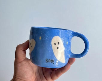 May include: Blue ceramic mug with a handle, decorated with white ghost illustrations and yellow star accents. The word "boo!" is written in black. The mug is held by a hand.