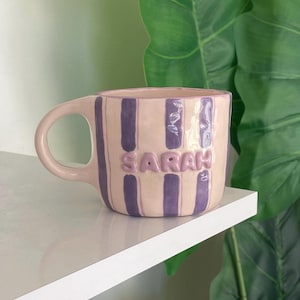 Può includere: Una tazza in ceramica con base rosa chiaro e strisce verticali viola. Il nome "Sarah" è scritto in lettere in rilievo. La tazza ha un manico rosa abbinato ed è esposta su una mensola bianca.