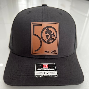 May include: Black trucker hat with a brown leather patch featuring a "50" graphic, Mickey Mouse, and the dates "1971-2021." The hat includes a Richardson tag with "Original 112 Trucker" text.