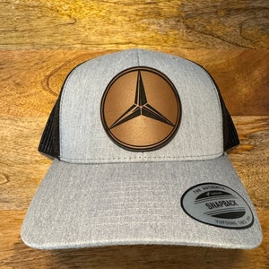 May include: A gray and black trucker hat with a brown leather patch featuring a three-pointed star logo. The hat has a black mesh back and a gray front with a black adjustable strap. The text "The Authentic Snapback" is printed on a black tag attached to the strap.