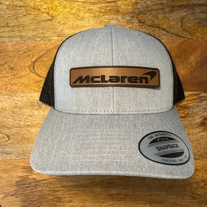 May include: A gray and black baseball cap with a brown leather patch that says "McLaren" on the front. The cap has a black mesh back and a black "Yupoong The Authentic Snapback" label on the back.