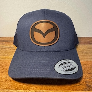 May include: A navy blue trucker hat with a brown leather patch featuring the Mazda logo. The hat has a mesh back and a black "The Authentic Yupoong Snapback Yupoong Since 1974" label.