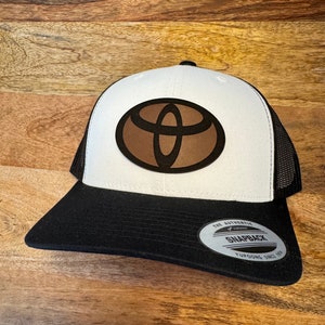 Könnte beinhalten: Weiße und schwarze Trucker-Kappe mit einem braunen Leder-Toyota-Logo. Die Kappe hat ein schwarzes Netzrücken und ein schwarzes Schild. Die Kappe hat ein Etikett mit der Aufschrift "The Authentic Yupoong Snapback Yupoong Since 1974".