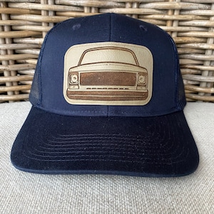 Puede incluir: Una gorra de camionero azul marino con un parche de cuero marrón que presenta un diseño de camión vintage.