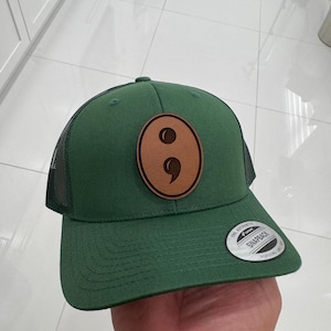 May include: A green trucker hat with a brown leather patch featuring a semicolon design. The hat has a mesh back and a curved brim. The hat has a snapback closure and a small white tag on the brim.