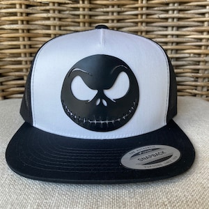 May include: Black and white trucker hat with a black Jack Skellington face on the front. The hat has a black mesh back and a black brim with a white "The Authentic Yupoong Snapback Yupoong Since 1974" sticker.