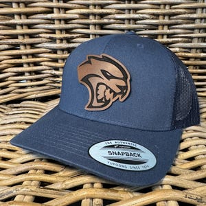 May include: A dark blue and black mesh baseball cap with a brown leather patch featuring a snarling animal head. The cap has a curved brim and a snap closure. The label on the cap reads "The Authentic Yupoong Snapback Yupoong Since 1974".