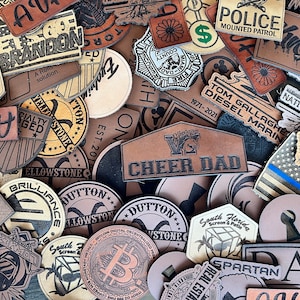 May include: A collection of brown leather patches with various designs and text, including "Cheer Dad", "Yellowstone", "Dutton", "South Florida Screen & Patio", "Spartan", "Native Roots", "Police Mounted Patrol", and "Tom Gallagher Diesel Marine".