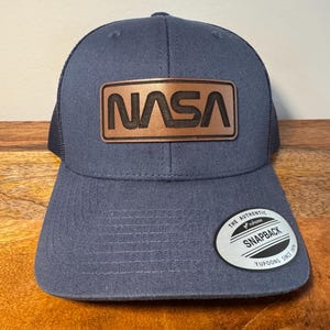 May include: A navy blue trucker hat with a brown leather patch that says "NASA" in black lettering. The hat has a black mesh back and a white sticker that says "The Authentic Yupoong Snapback Since 1974".