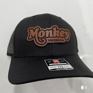 May include: Black mesh trucker hat with a brown leather patch that says "Monkey Honda" in a retro font. The hat is adjustable and has a Richardson logo on the side.