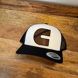 May include: A black and white trucker hat with the word "Cummins" in brown lettering. The hat has a white front panel, a black brim, and a black mesh back. The hat is sitting on a wooden surface.