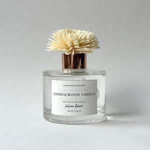 Könnte beinhalten: Eine klare Glasflasche mit einem weißen Etikett, auf dem "Luxury Reed Diffuser Sandalwood Vanilla Exclusive for you by Laluna Decor 200 ml / 6.7 fl oz" steht. Die Flasche hat einen weißen, blumenförmigen Diffusor oben drauf.