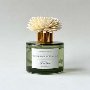 Può includere: Una bottiglia di vetro verde con un tappo dorato e un fiore bianco in cima. La bottiglia contiene un diffusore di canne con l'etichetta "LUXURY REED DIFFUSER WOOD SAGE & SEA SALT EXCLUSIVE FOR YOU BY Laluna Decor 100 ml / 3.5 fl oz".