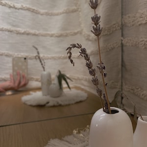 May include: A white ceramic vase contains dried lavender stems. The vase sits on a wooden surface, with a mirror reflecting the vase and other decorative items. A white macrame placemat is in the foreground.