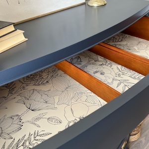 K&ouml;nnte beinhalten: Eine dunkelblaue Kommode mit einer geschwungenen Front und drei Schubladen. Die Schubladen sind mit einem wei&szlig;en Blumenmuster ausgekleidet.