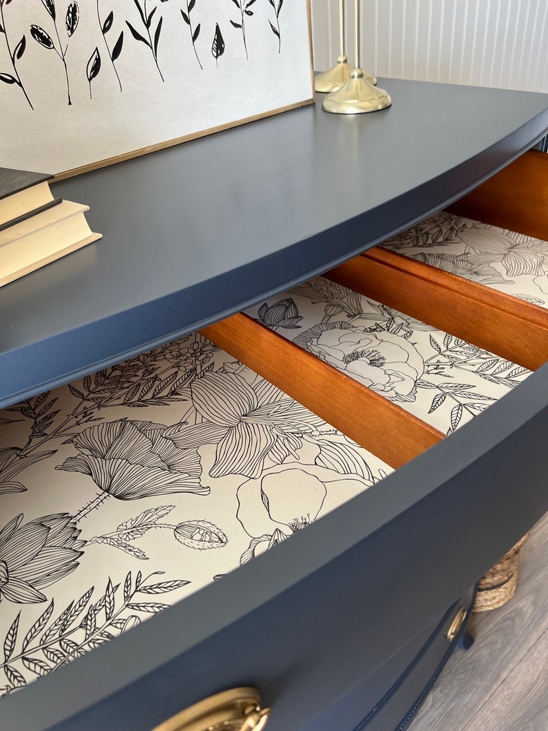 K&ouml;nnte beinhalten: Eine dunkelblaue Kommode mit einer geschwungenen Front und drei Schubladen. Die Schubladen sind mit einem wei&szlig;en Blumenmuster ausgekleidet.