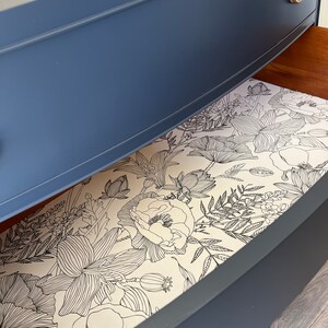 K&ouml;nnte beinhalten: Eine blaue Kommodenschublade mit einem floralen Futter. Das Futter ist ein wei&szlig;er Hintergrund mit schwarzen Linienzeichnungen von Blumen und Bl&auml;ttern.