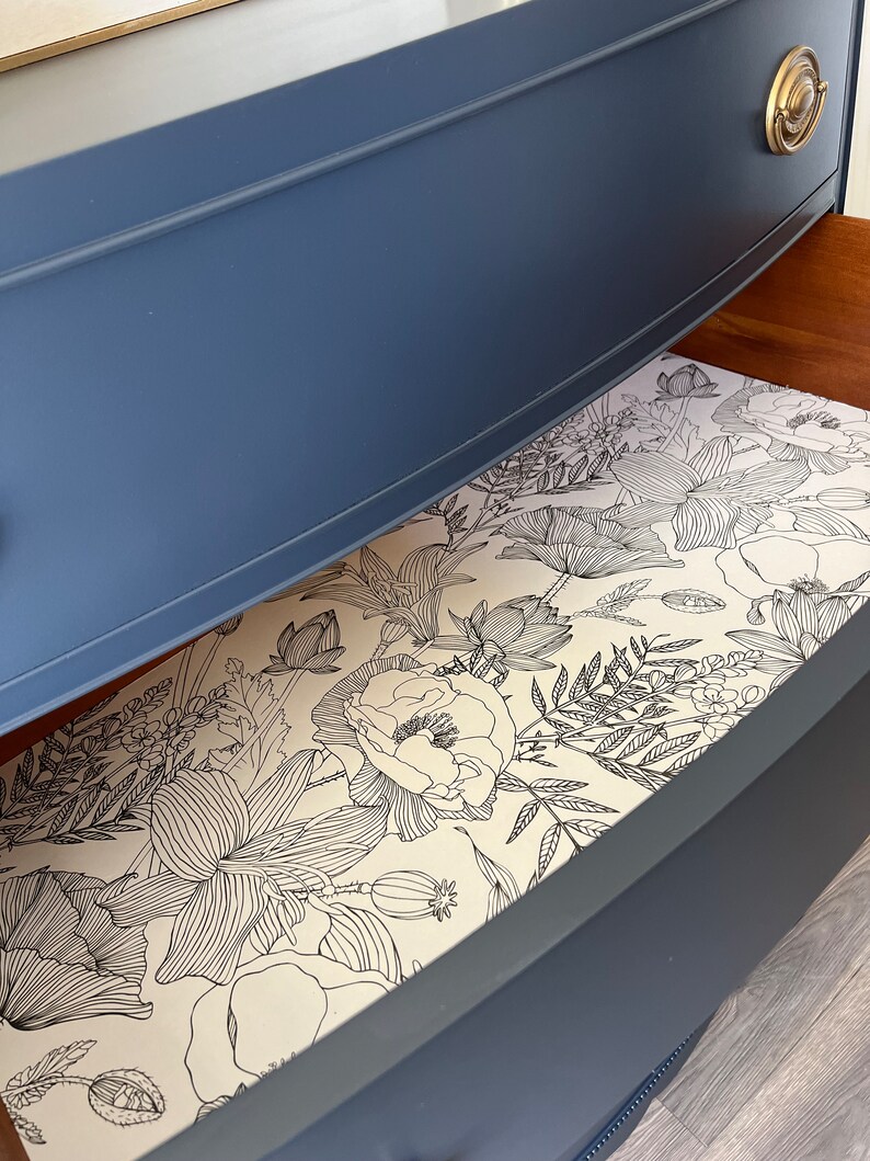 K&ouml;nnte beinhalten: Eine blaue Kommodenschublade mit einem floralen Futter. Das Futter ist ein wei&szlig;er Hintergrund mit schwarzen Linienzeichnungen von Blumen und Bl&auml;ttern.