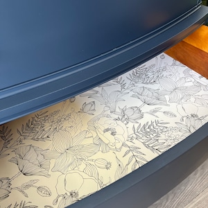 K&ouml;nnte beinhalten: Eine blaue Kommodenschublade mit einem floralen Futter. Das Futter ist ein wei&szlig;er Hintergrund mit schwarzen Linienzeichnungen von Blumen und Bl&auml;ttern.