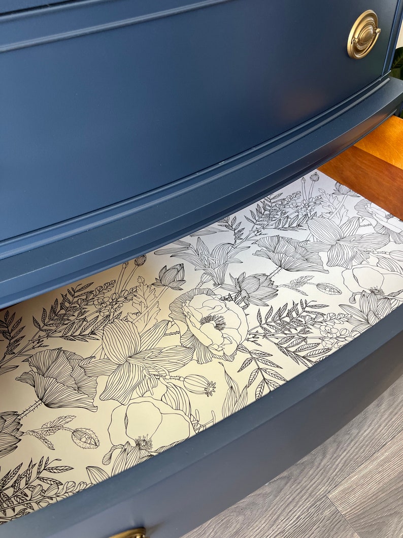 K&ouml;nnte beinhalten: Eine blaue Kommodenschublade mit einem floralen Futter. Das Futter ist ein wei&szlig;er Hintergrund mit schwarzen Linienzeichnungen von Blumen und Bl&auml;ttern.
