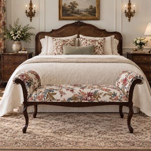 May include: A wooden bench with curved arms and legs, upholstered in a cream fabric featuring a multi-colored floral pattern. The bench is positioned in front of a bed with a dark wood headboard, white bedding, and floral accent pillows.