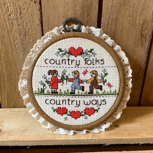 May include: A framed cross-stitch with a white background, brown wooden frame, and white lace trim. The cross-stitch features three children holding hands and walking, with the text "country folks" above them and "country ways" below them. There are red hearts and blue flowers throughout the design.