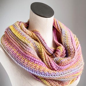 Könnte beinhalten: Ein grob gestrickter Infinity-Schal mit einem melierten Garnmuster in den Farben zartes Rosa, sonniges Gelb, Lavendel und Creme. Der Schal ist in einem strukturierten Rippenmuster gefertigt, das für ein dickes, mehrschichtiges Erscheinungsbild sorgt.