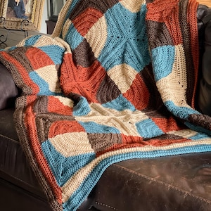 Peut inclure: Une couverture crochetée à la main avec un motif patchwork, composée de carrés rouille, marron, beige et bleu sarcelle. La couverture est drapée sur un canapé en cuir marron, avec un pompon bleu sarcelle au coin. Un élément de décoration intérieure douillet.