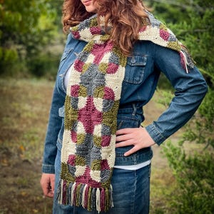 May include: A handmade crochet scarf with a geometric pattern in shades of cream, burgundy, gray, and olive green. The scarf has a fringed edge and is worn over a denim jacket. The scarf is a cozy accessory.