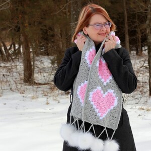 May include: A gray and pink crocheted scarf with heart-shaped designs. The scarf has a white fur pom-pom on each end.
